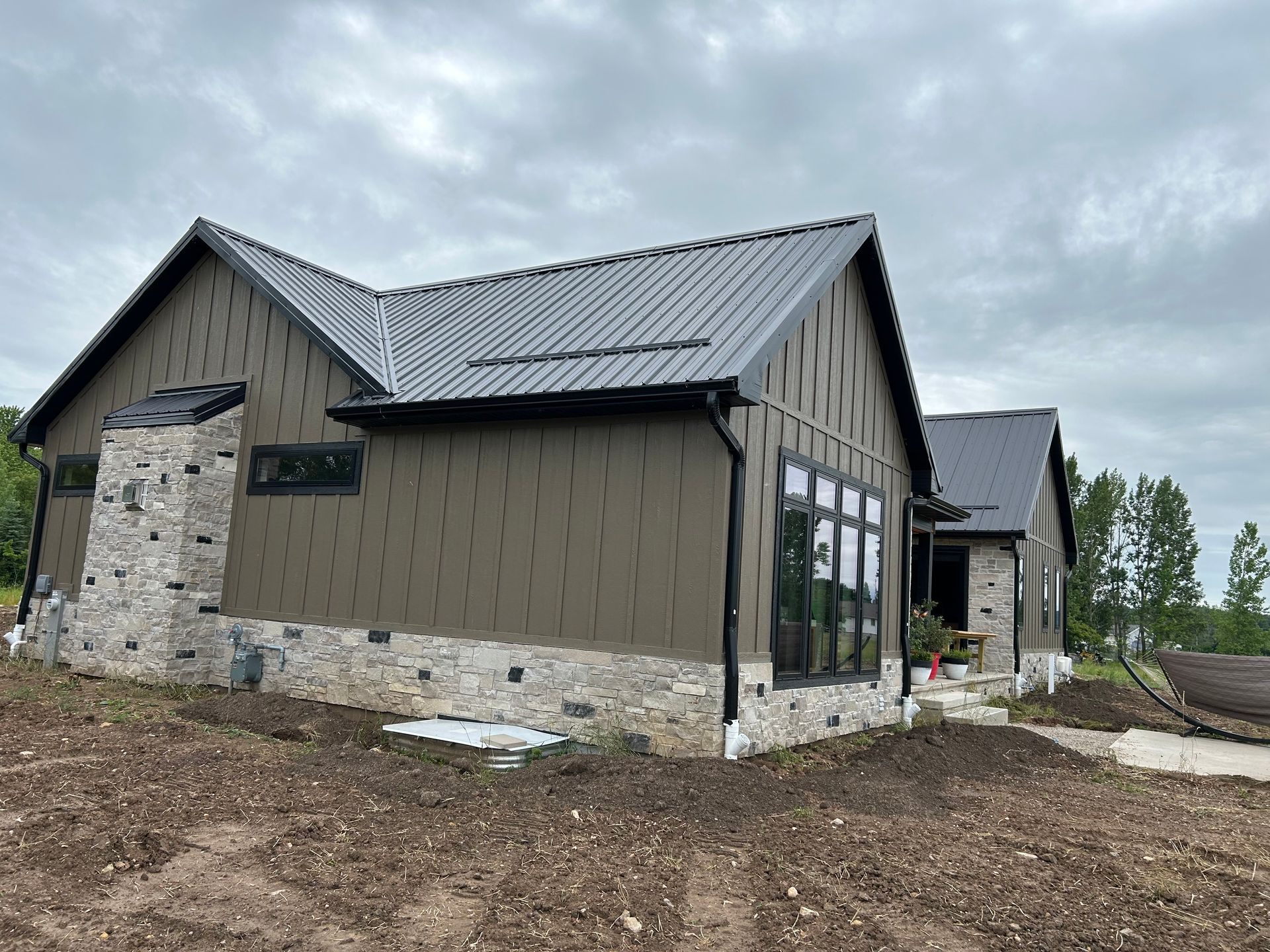 Modern house with olive-brown siding, stone accents, and dark metal roof under a cloudy sky.