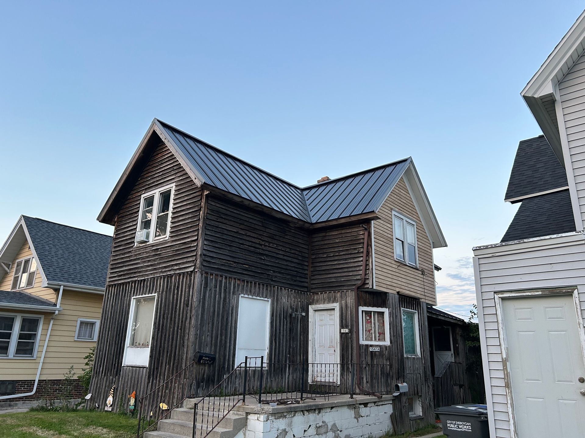Two-story weathered house with dark siding and metal roof; next to a light-colored house under a blue sky.