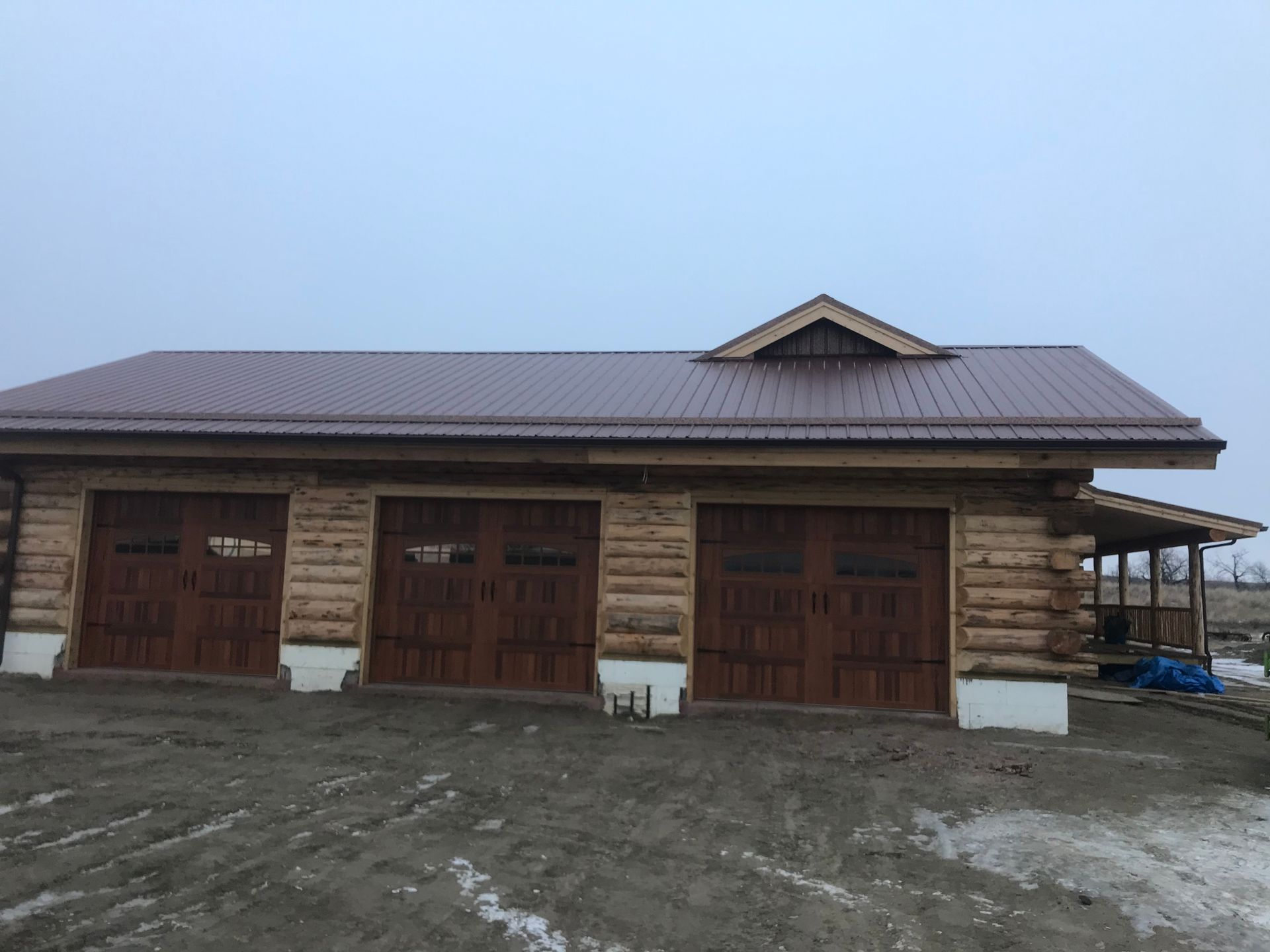 Brown log cabin garage with three doors, brown roof, and small porch. Cloudy, outdoor setting.