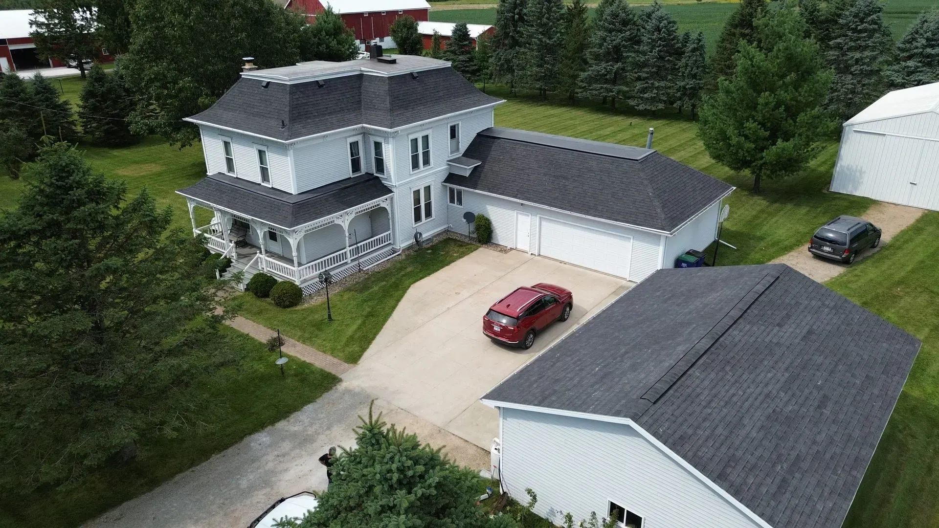 An aerial view of a white farmhouse with a dark roof, a connected garage, a detached building, and cars in the driveway