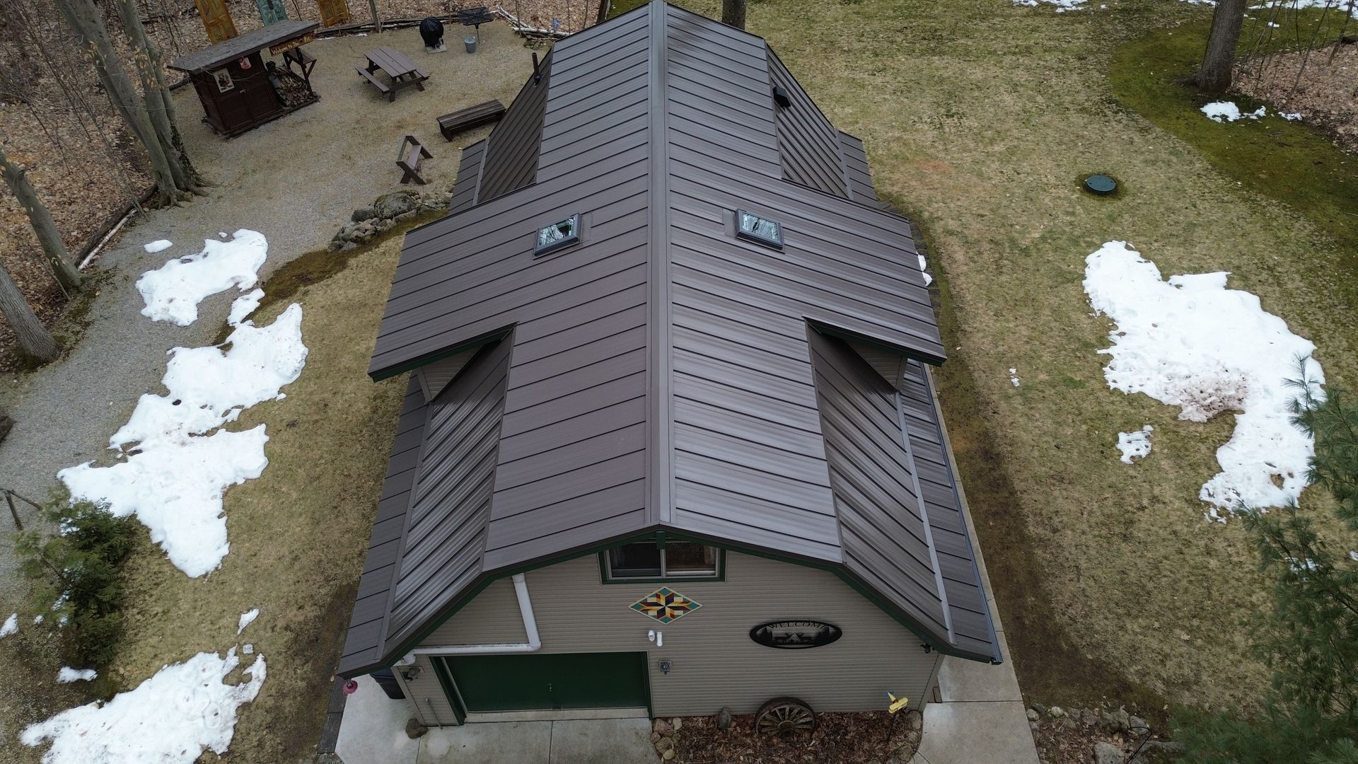An aerial view of a gray building with a dark brown metal roof, set on a grassy, partially snow-covered lot.