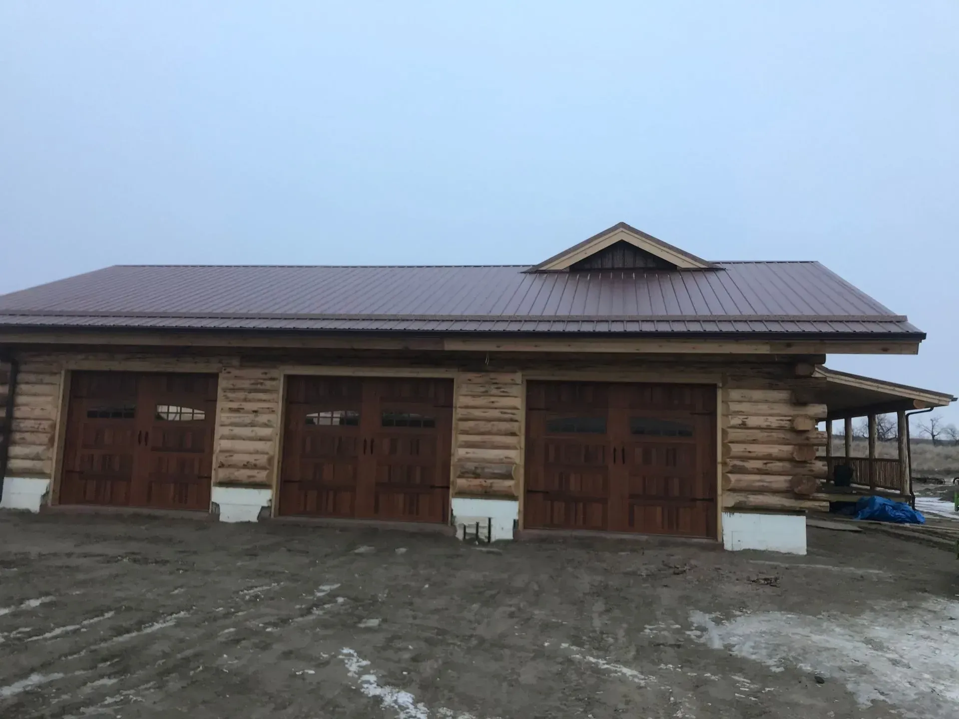 A log cabin-style garage with three wooden carriage-style doors and a dark metal roof under a gray, overcast sky