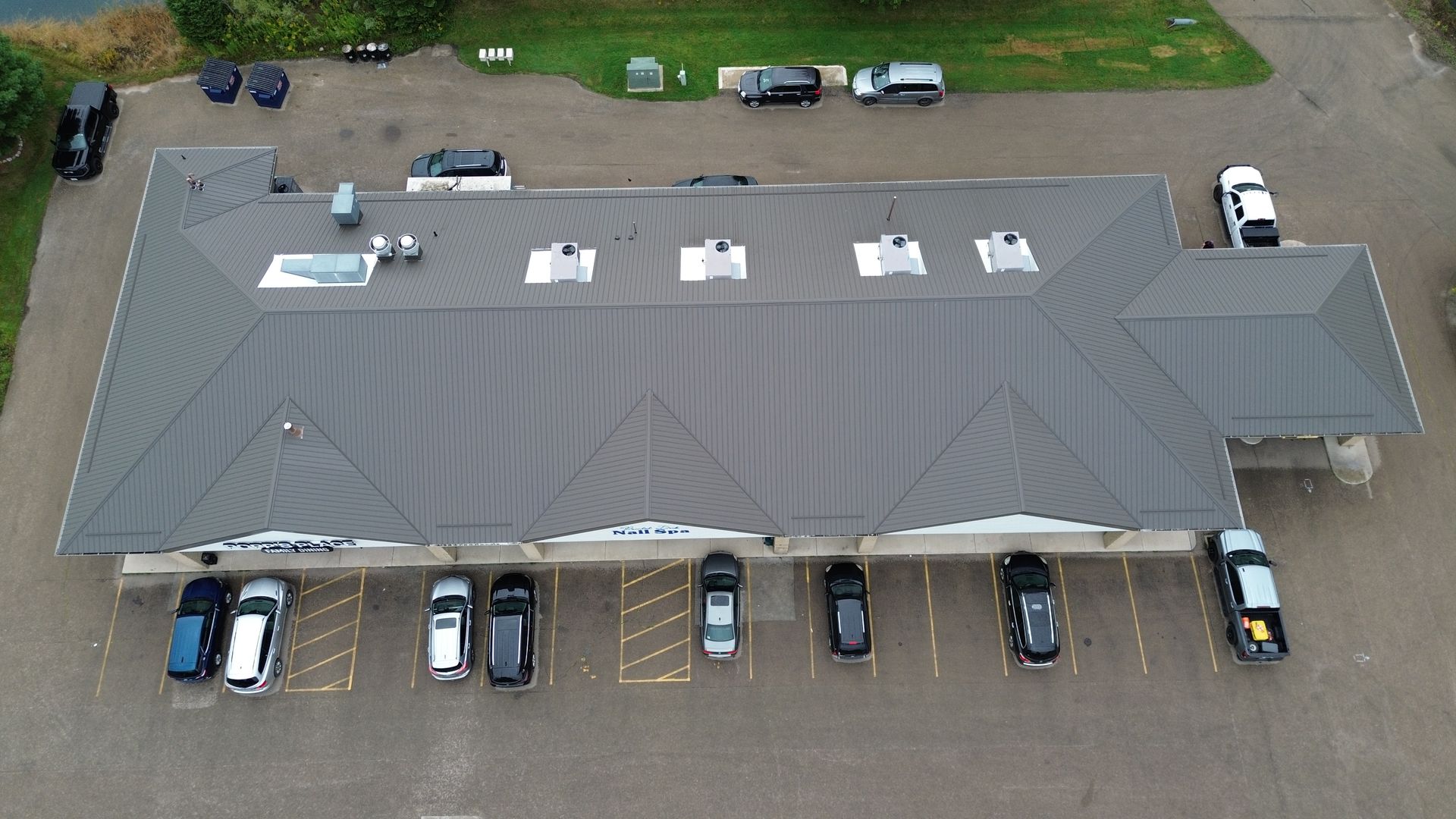 Aerial view of a gray-roofed building with parked cars around it and a gravel lot.