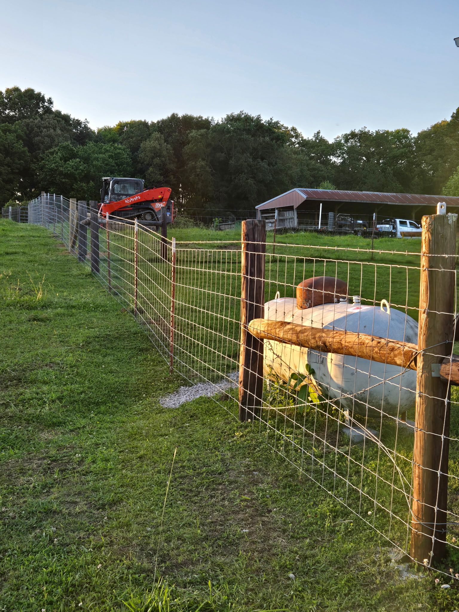 Farm with a fence, an excavator, a propane tank, and a shed in the background. Green grass and trees under a blue sky.