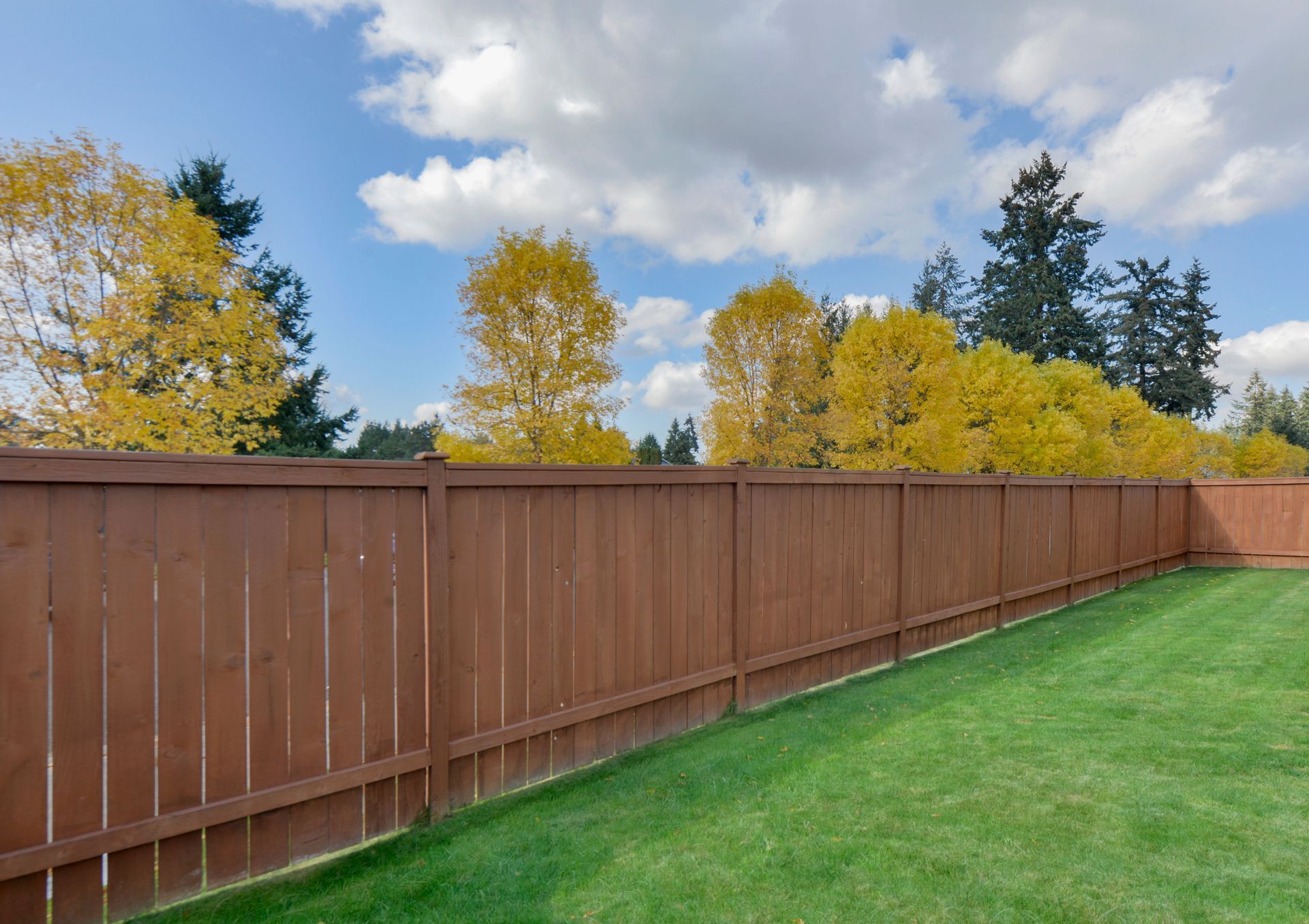 Brown wooden fence bordering a green lawn with trees and a blue sky with clouds.