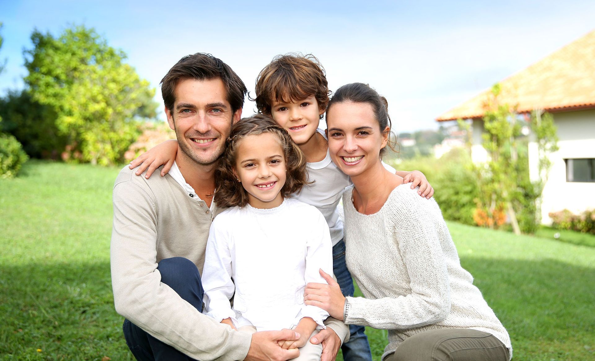 Family of four smiling in a sunny yard; parents embrace two children.