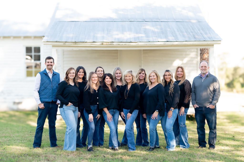 Group of people in front of white building. Blue jeans, black tops, smiling, outdoor setting.