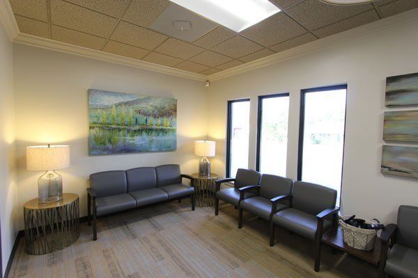 Waiting room with gray seating, artwork, lamps, and windows; neutral colors.