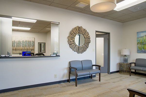 Waiting room with gray chairs, a round mirror, and a reception window.