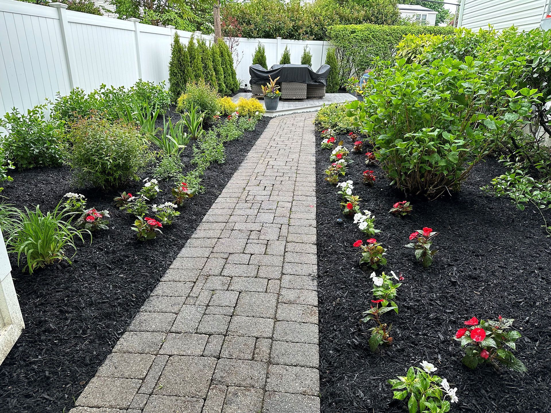 A brick walkway leading to a patio surrounded by flowers and bushes