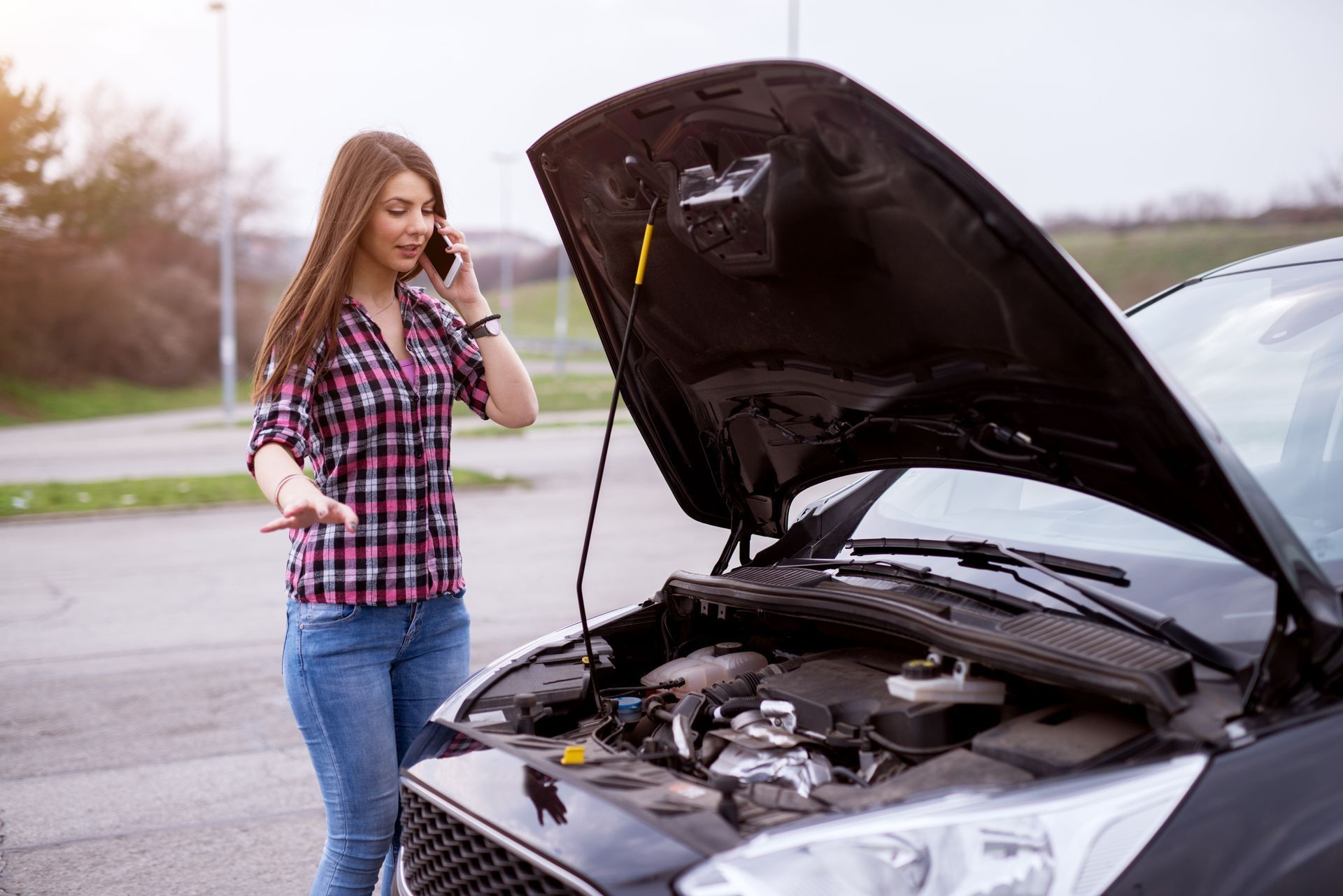 Woman standing beside a car with its hood open on a roadside, looking at the engine and holding a phone.