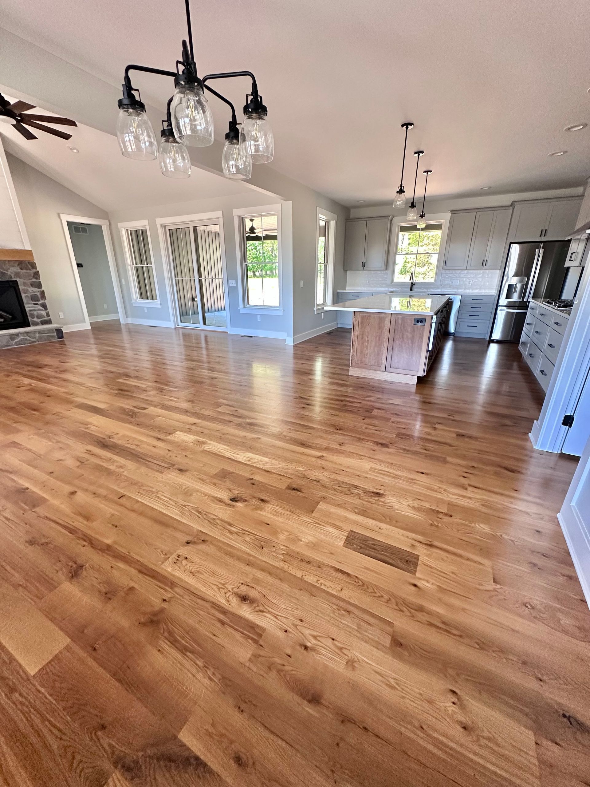 Bright open living room with hardwood floors, white walls, and a kitchen in the background.