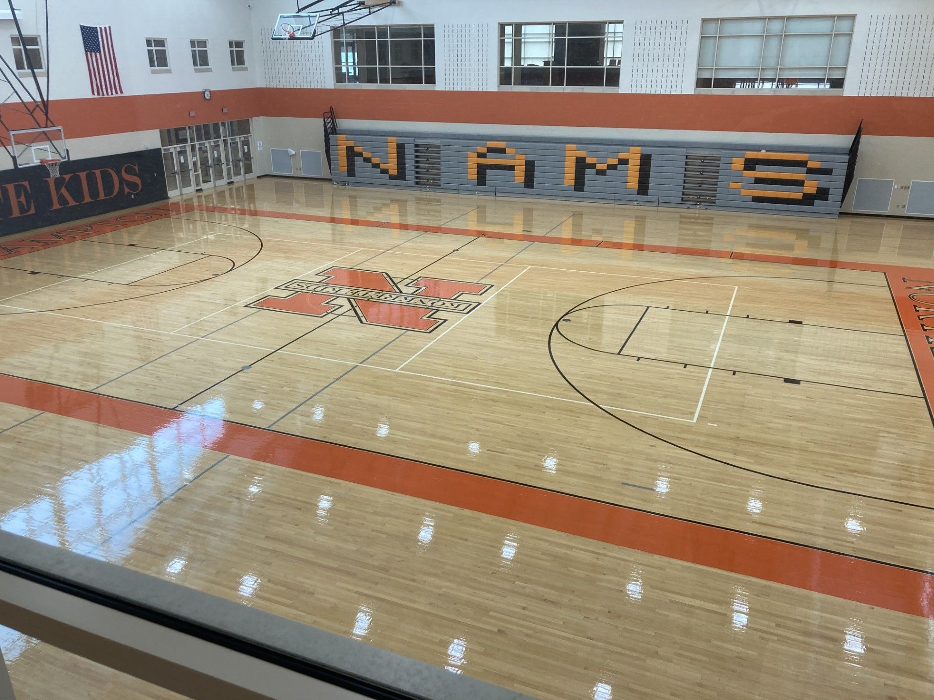 A high-angle view of a school basketball court with light wood floors, orange accents, and the school name NAMS on a wall.