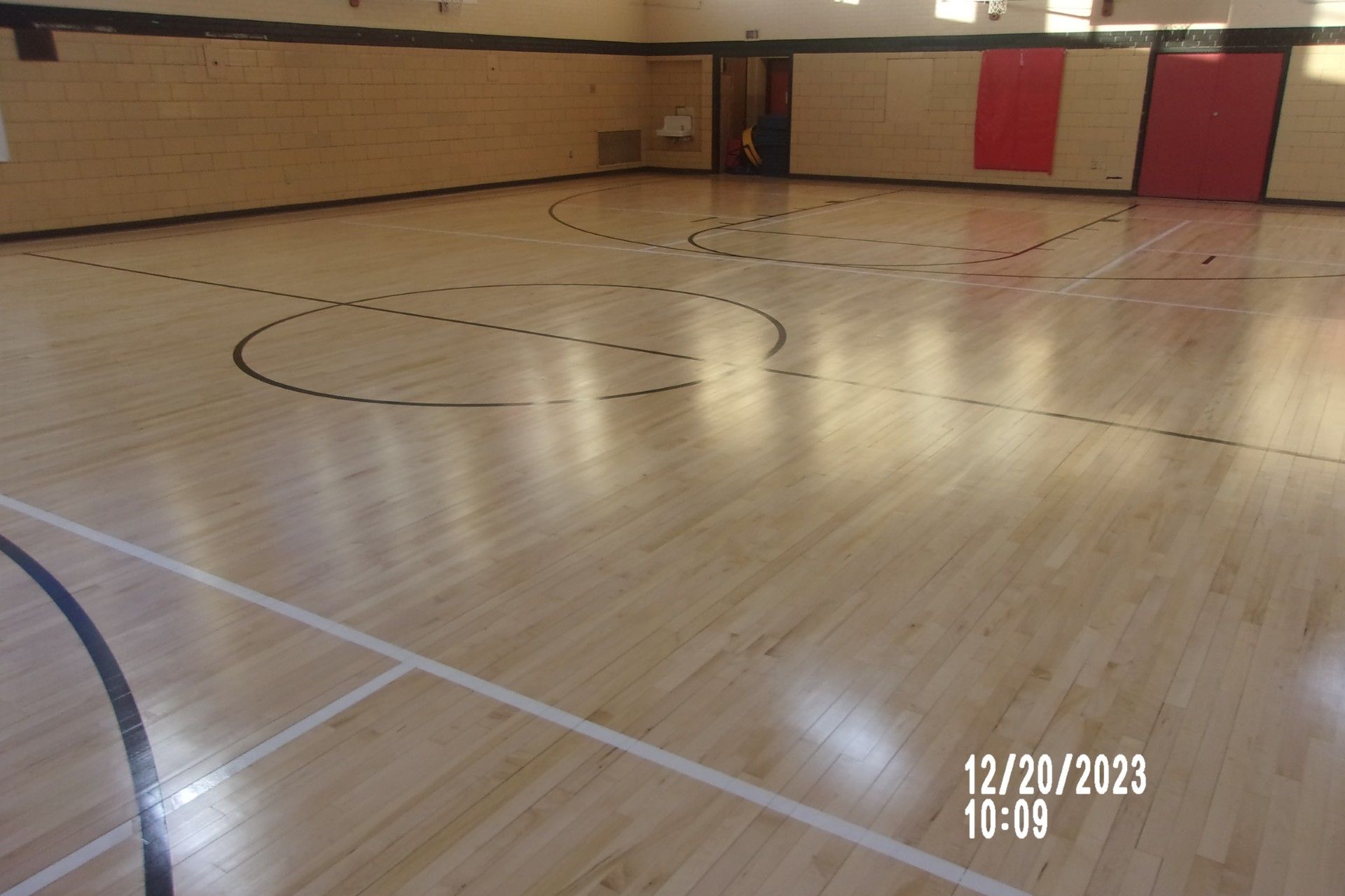 Empty gymnasium with polished wooden floor and court lines, red wall mats, and basketball hoops in the distance.