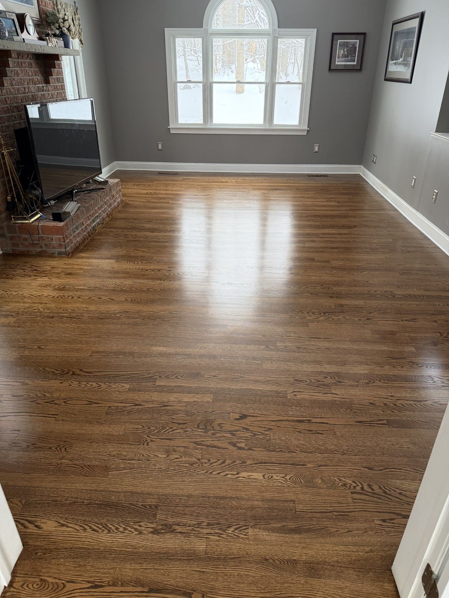 A sunlit room with polished hardwood floors, grey walls, and a large arched window overlooking a snowy exterior.