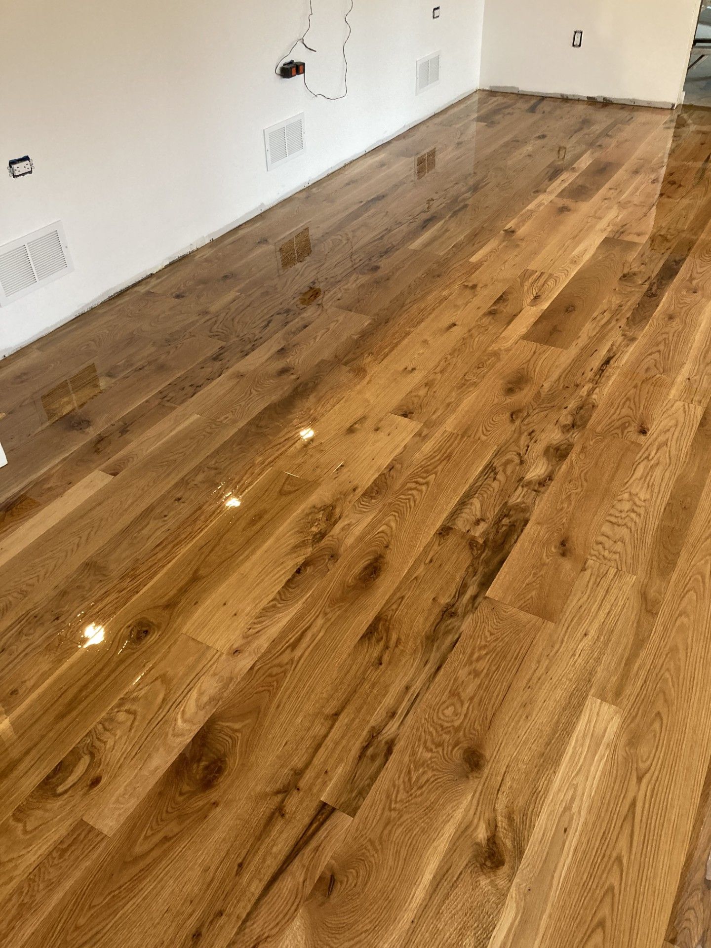Newly installed, glossy, light brown hardwood floor planks stretching towards a white wall with electrical outlets.