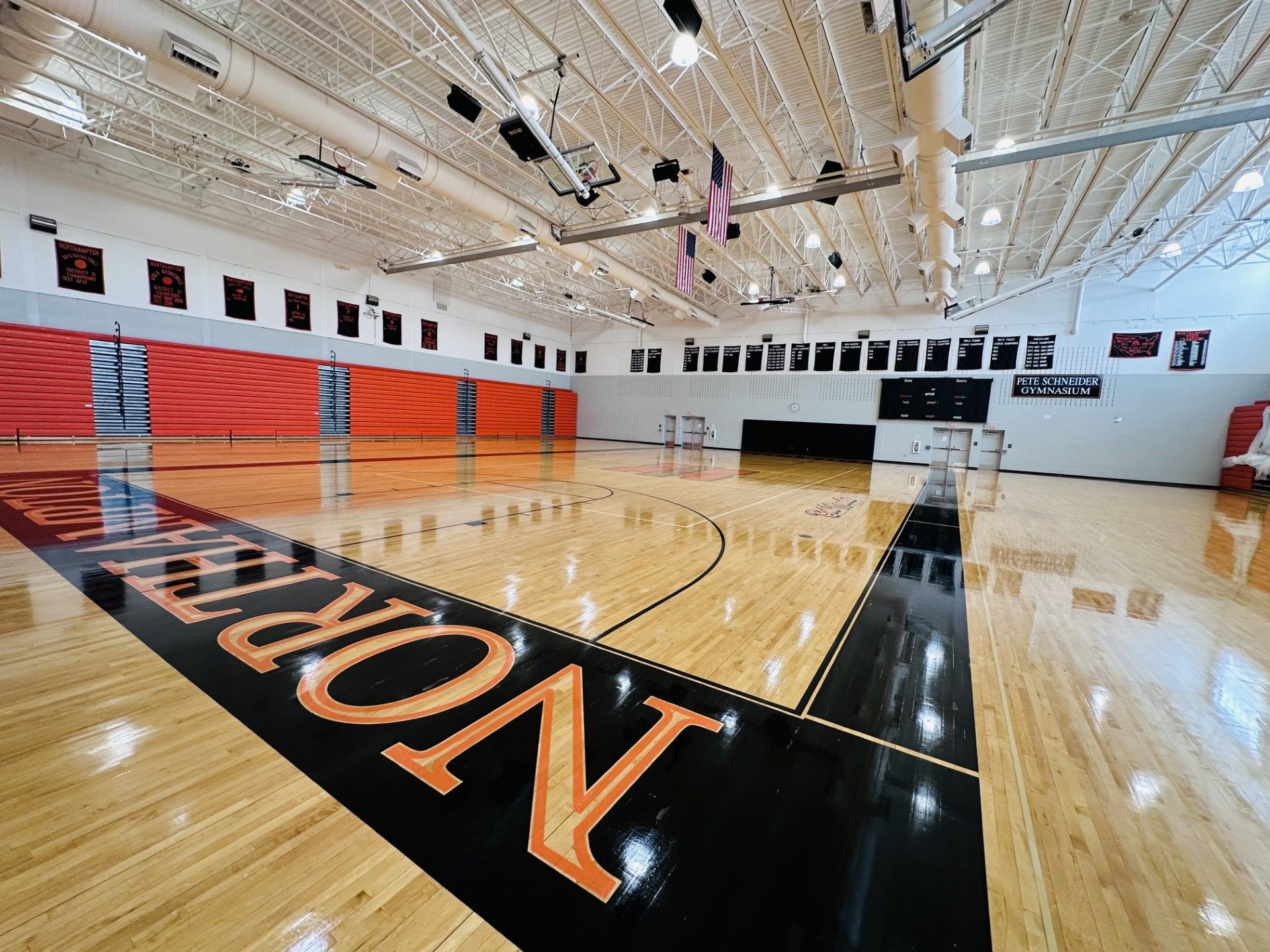 Empty gymnasium with polished wooden floor and court lines, red wall mats, and basketball hoops in the distance.