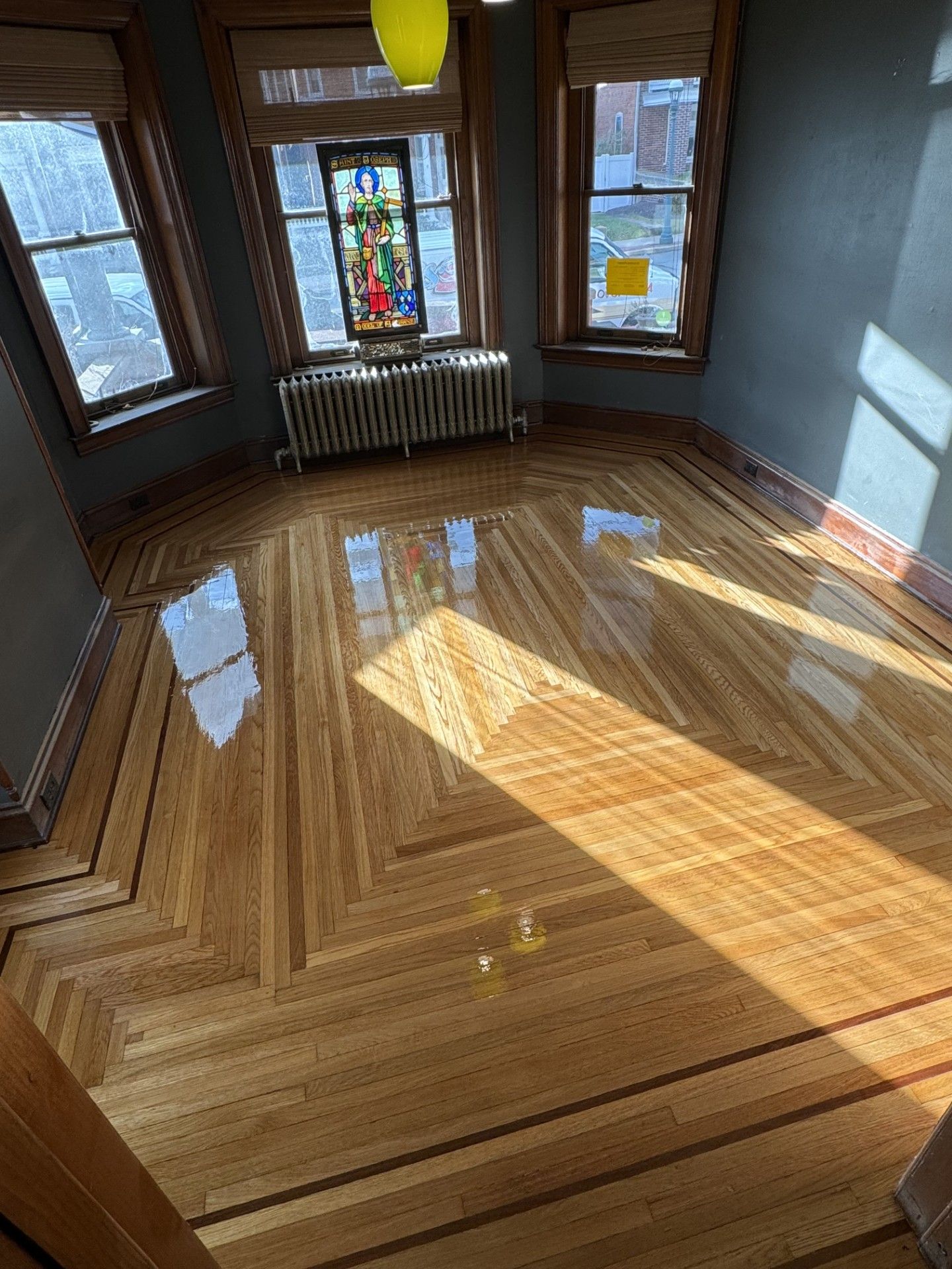 Sunlight illuminates polished wood flooring with a decorative geometric border in a room with large windows and a radiator.