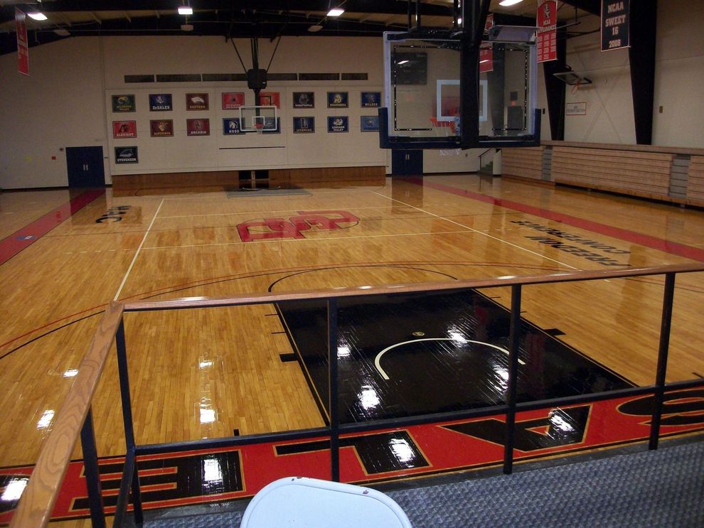 Basketball court, hardwood floor, black and red accents, empty bleachers, framed photos on the wall.