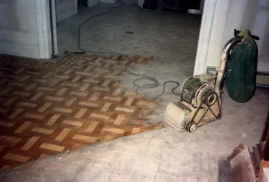 Floor sanding machine on parquet floor; half sanded, showing lighter wood.