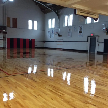 Empty basketball court with shiny wooden floor, black and red padded wall, and arched windows.