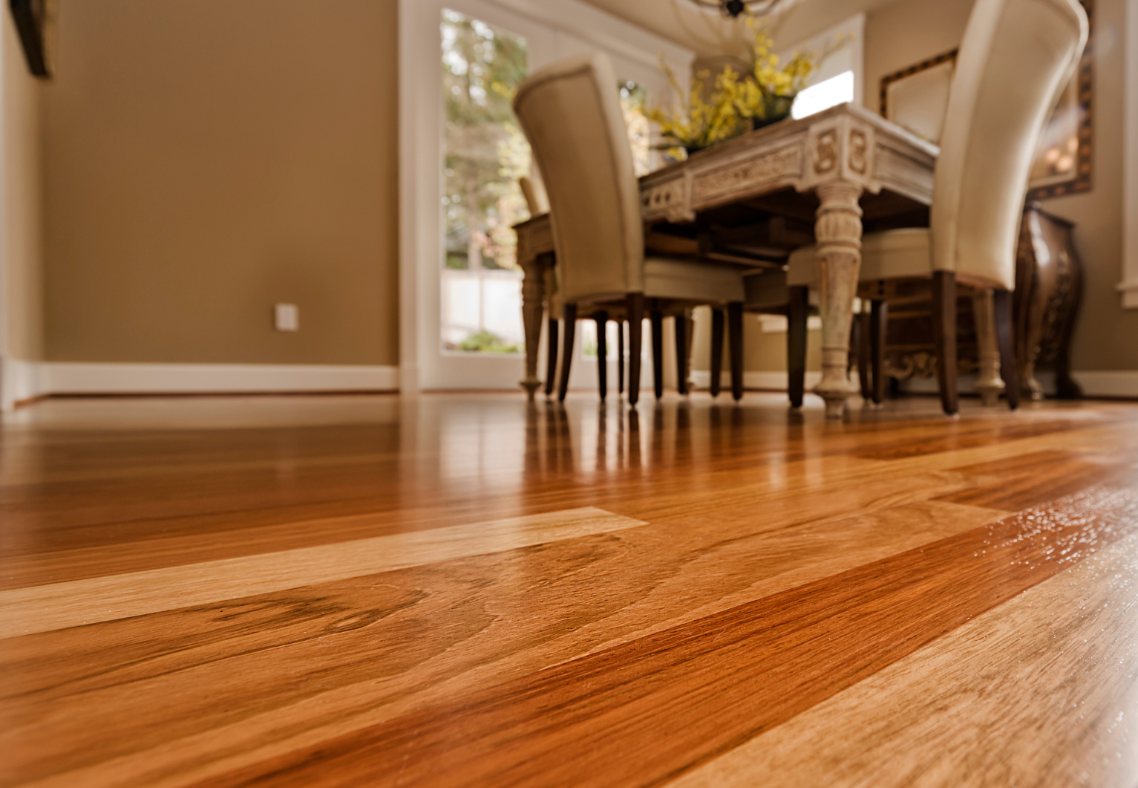 Close-up of polished hardwood flooring in a dining room; table and chairs visible in the background.