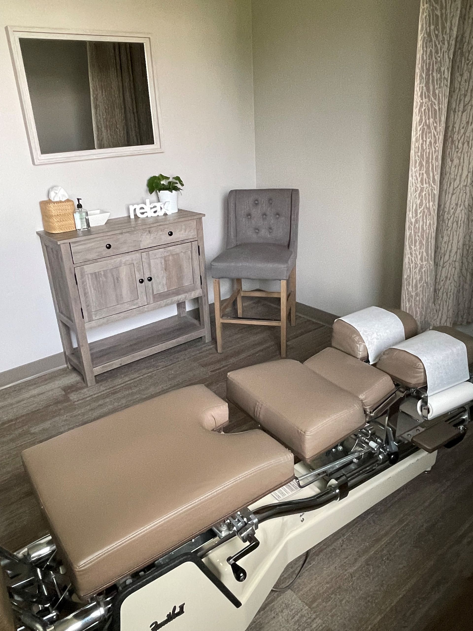 A room with a massage table, chair  mirror and dresser