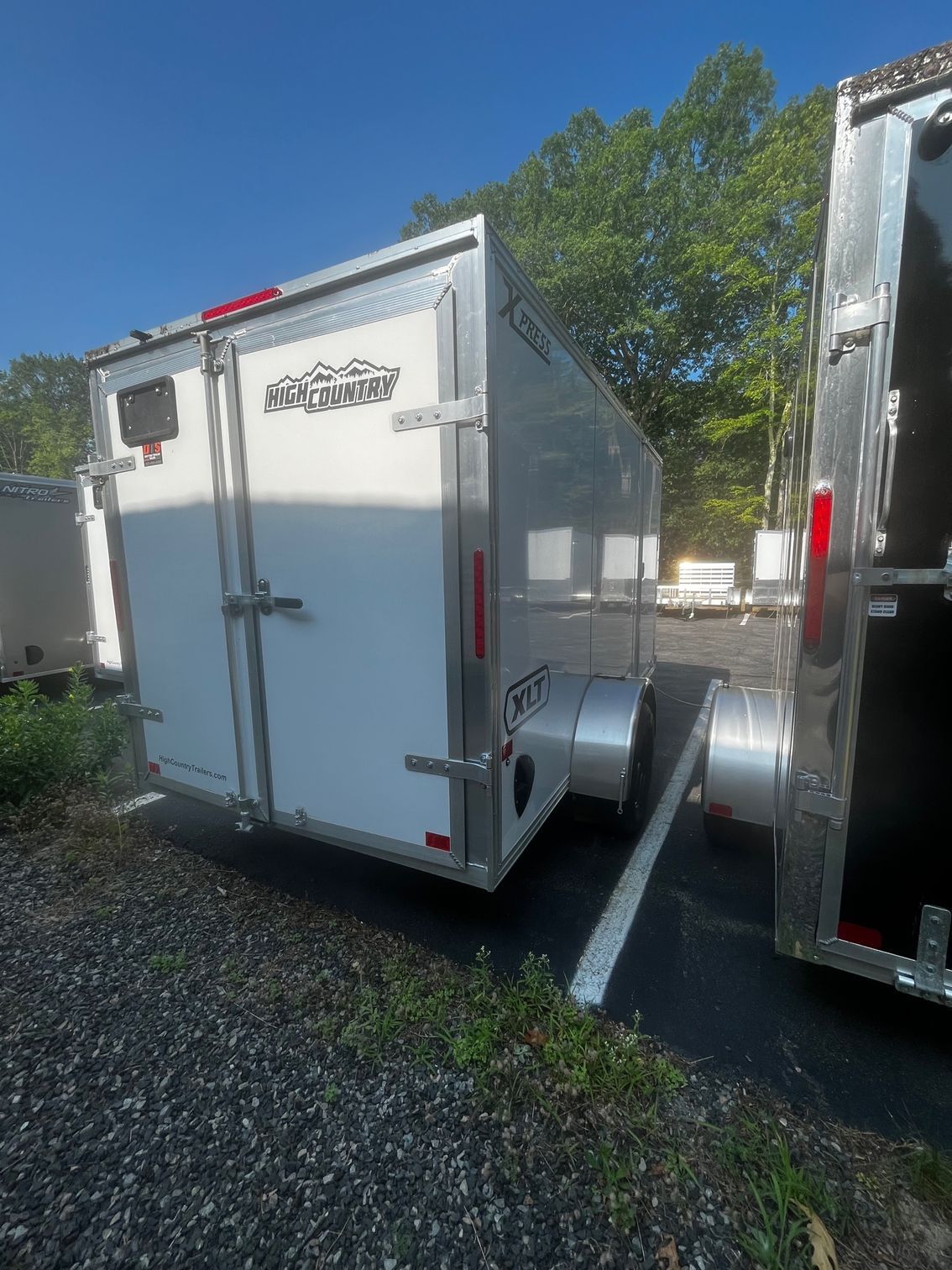 White and silver enclosed cargo trailer parked outside on asphalt, with a blue sky and trees in the background.