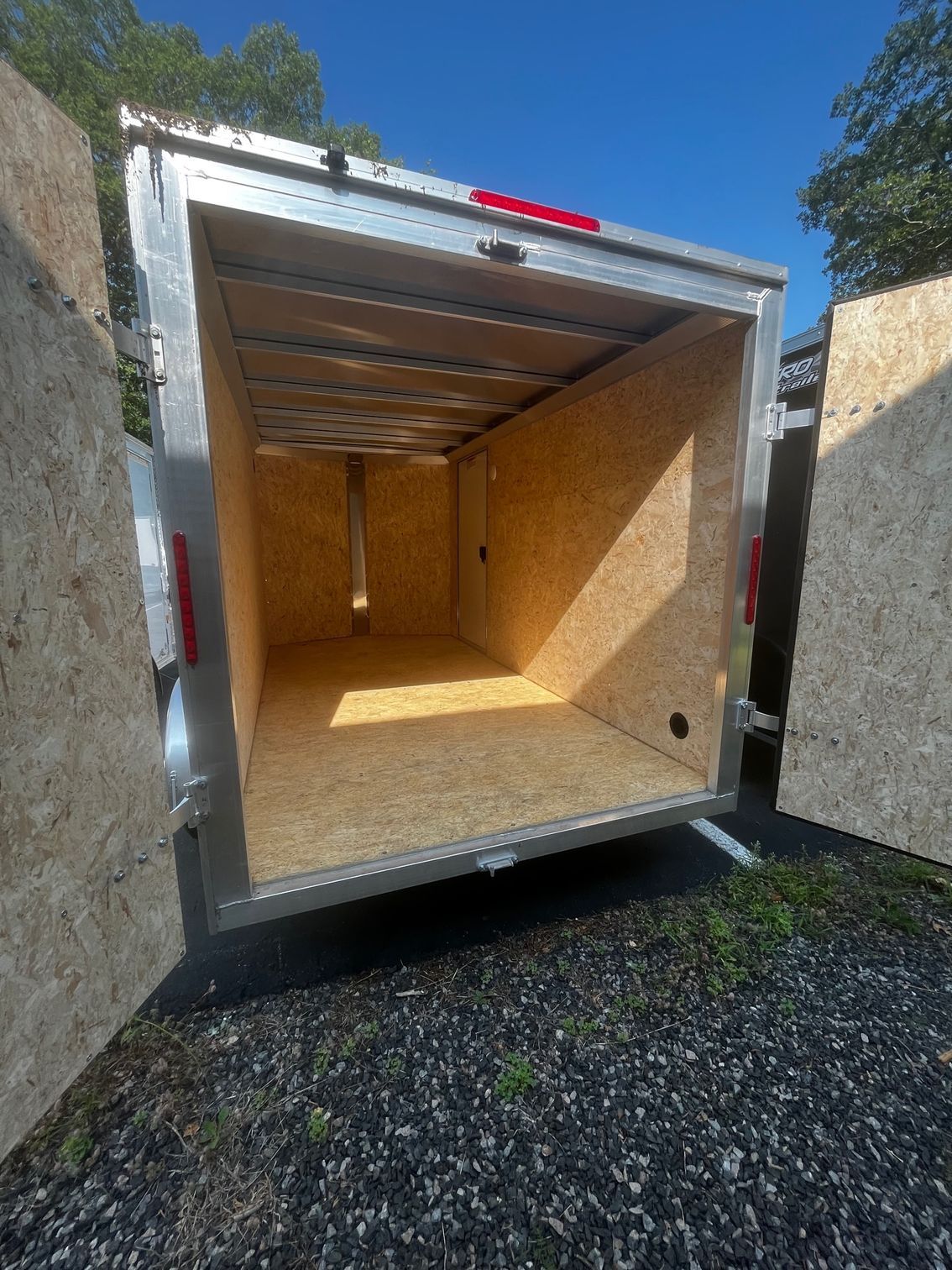 An open cargo trailer with wood paneling inside. The doors are open, revealing the interior. The trailer is parked outside.