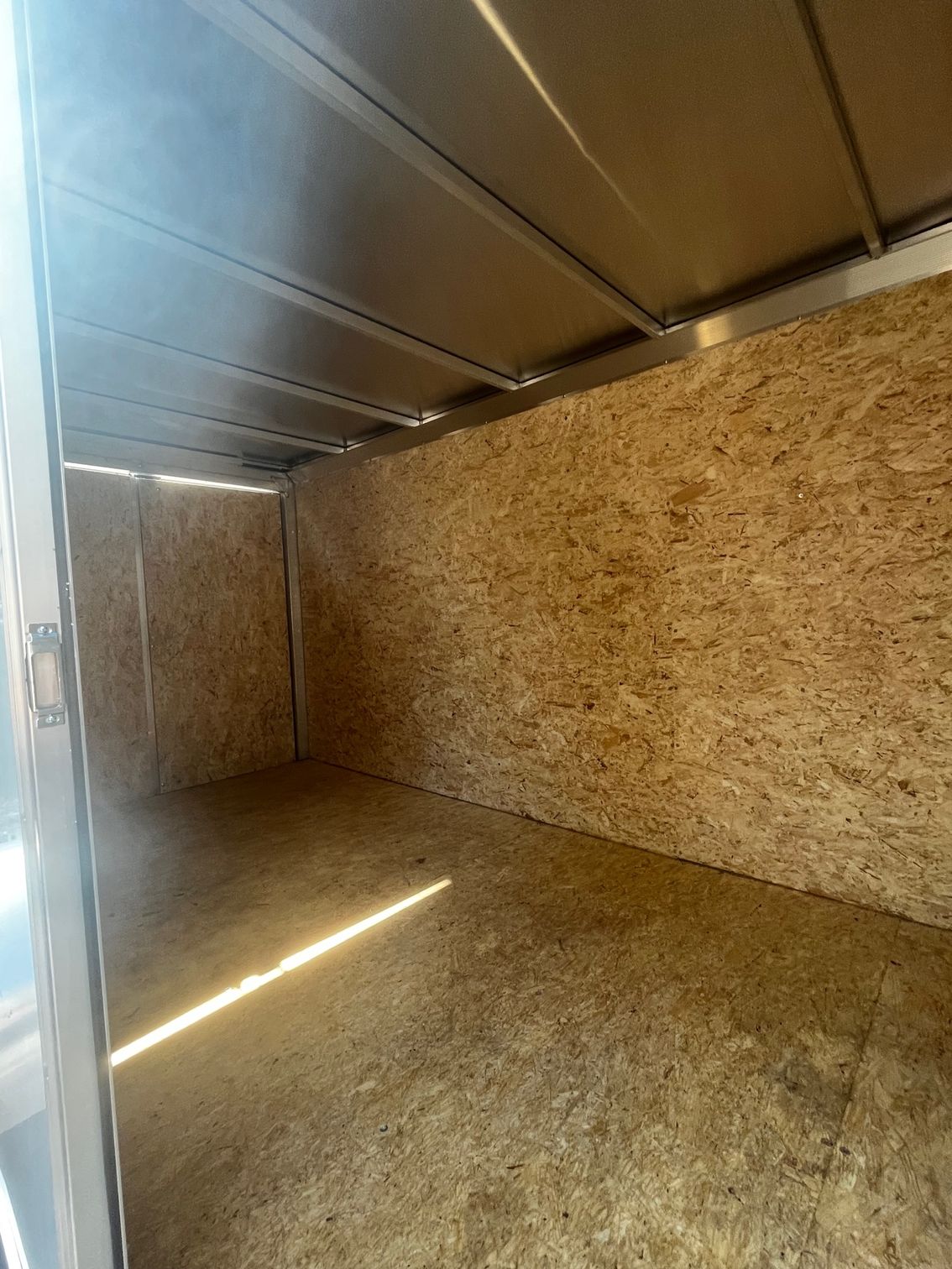 Empty cargo trailer interior with wood panel walls and floor, metallic ceiling, and open doorway.