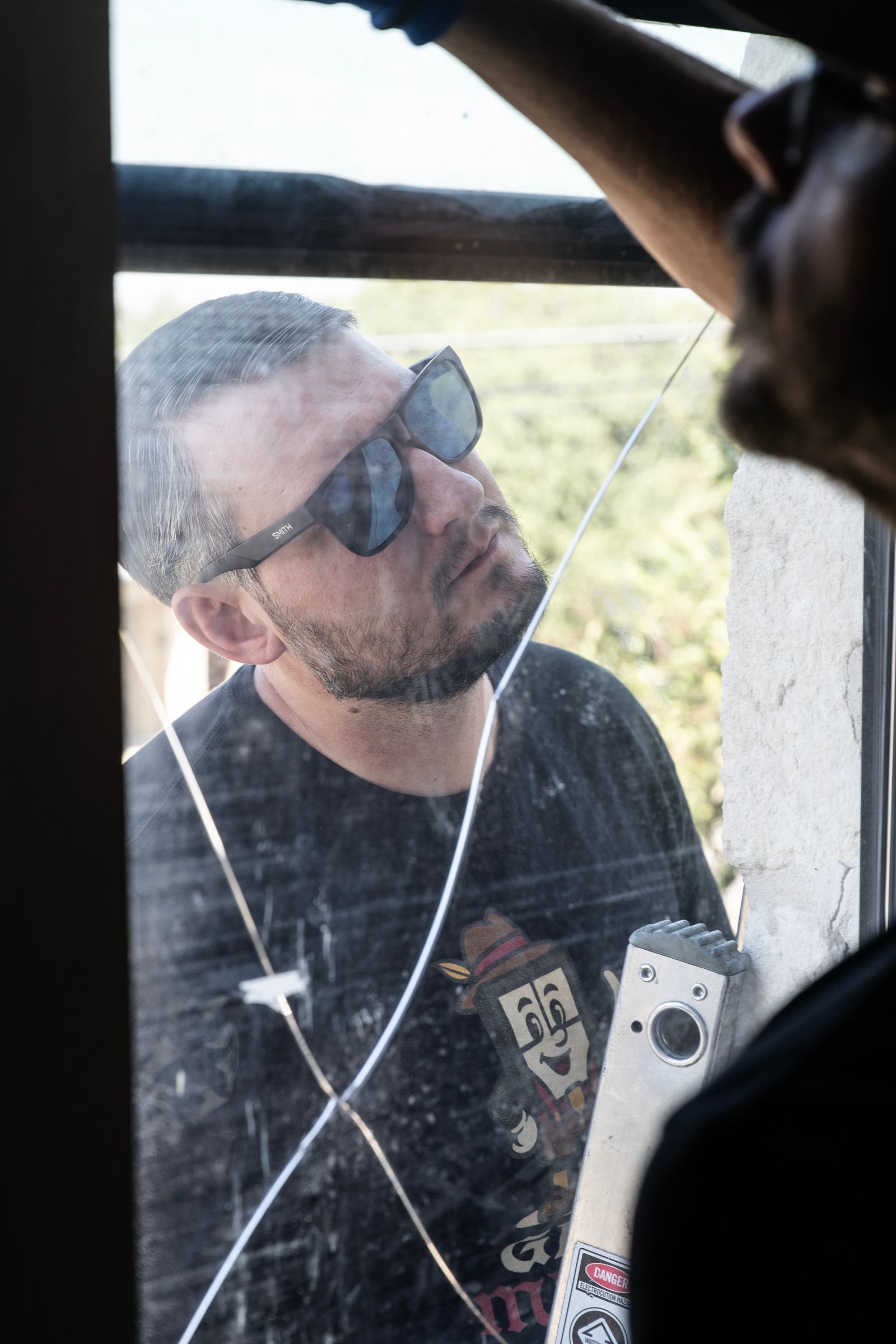 Man with sunglasses looking through a cracked window, another person visible on the right.