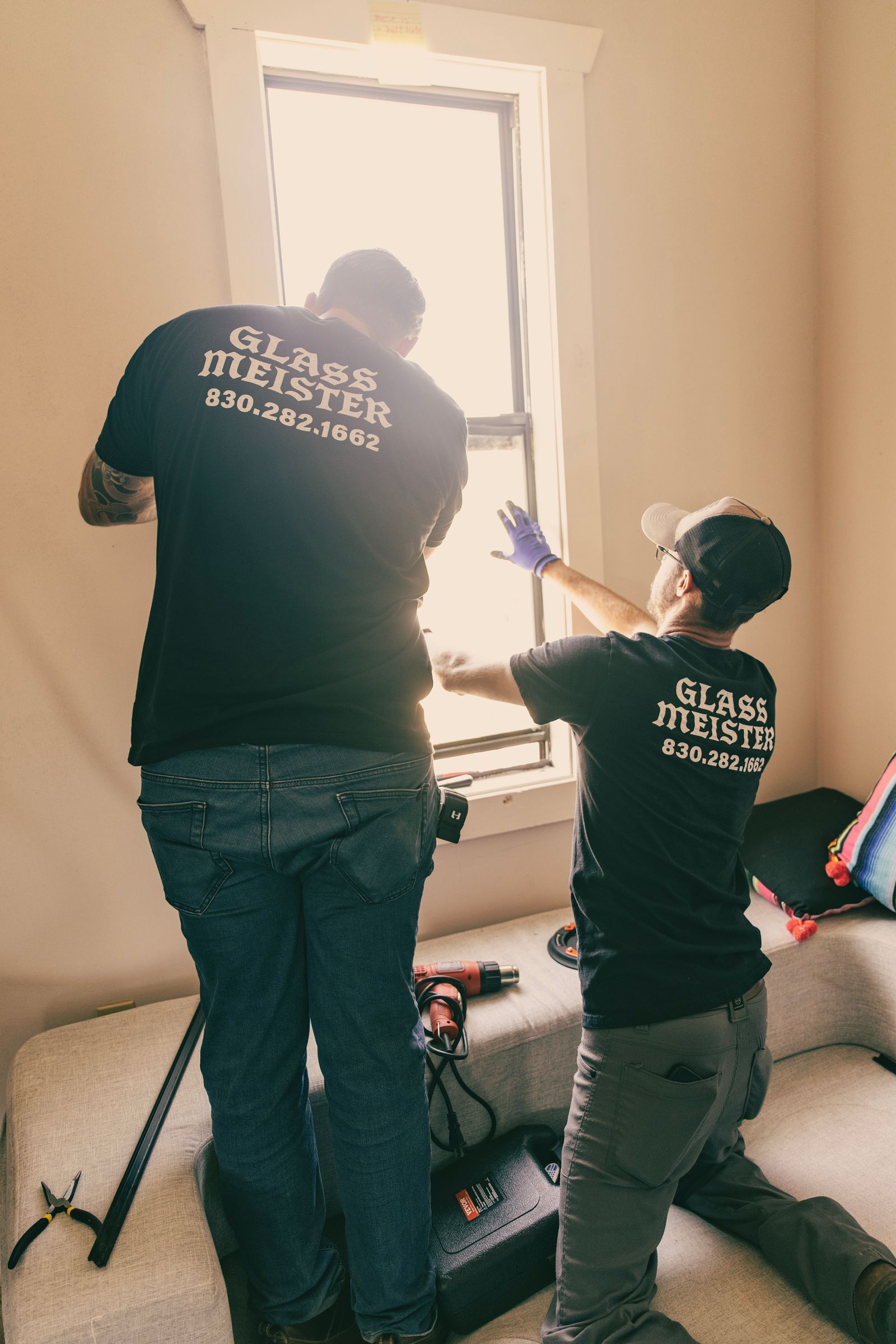Two people installing a window; they wear black shirts with company logo. Interior, bright light.