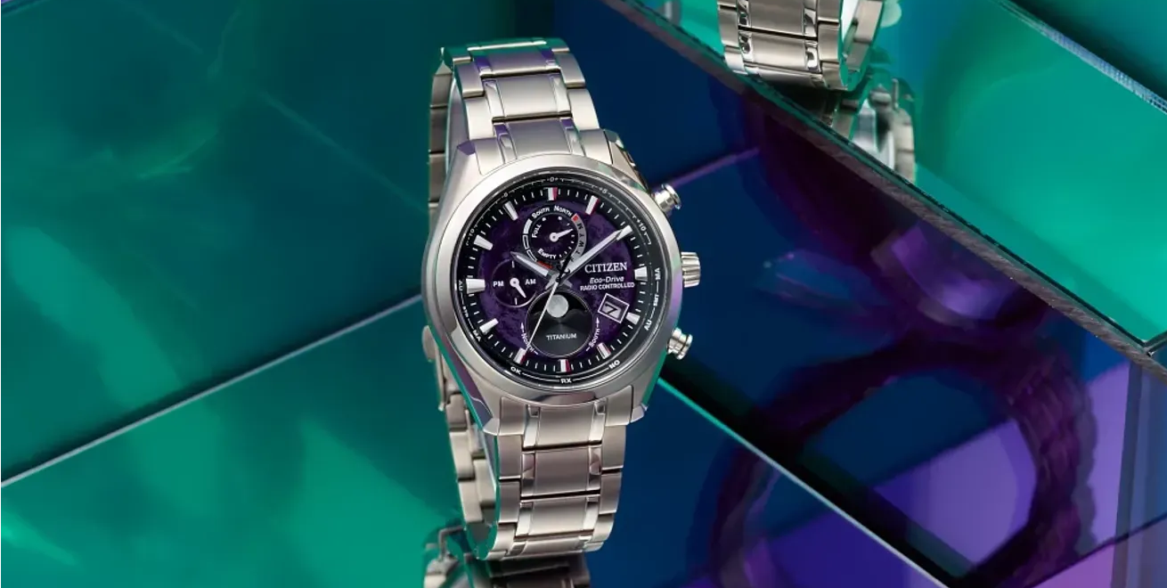 A silver-tone chronograph watch with a dark purple face, displayed on a reflective green and blue tiled surface.