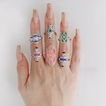 Hand displaying seven art deco style rings with various colored stones.