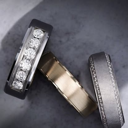 Three rings: one with diamonds in black setting, one gold, and one with diamonds in silver setting, on gray surface.