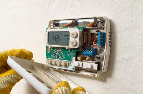 Person in gloves opening a thermostat on a white wall, revealing wires and controls.