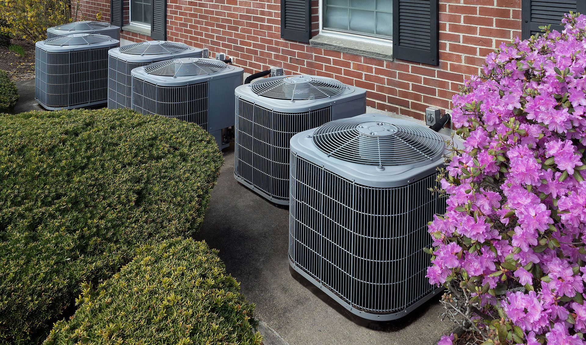 Four outdoor HVAC units beside a brick building, framed by green shrubs and pink flowers.
