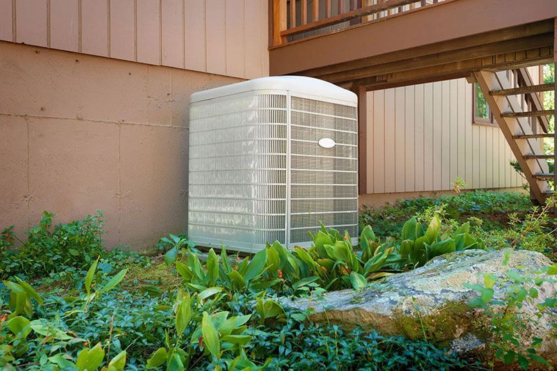 Air conditioning unit outside, beneath a wooden deck, surrounded by greenery.