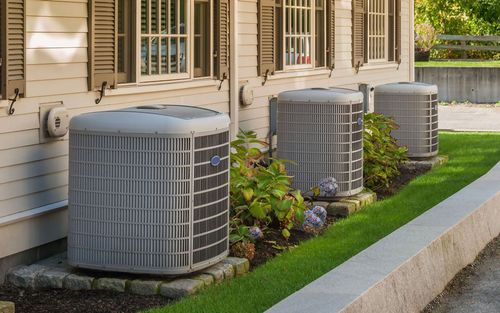 Three outdoor air conditioning units beside a beige house with shrubs and a concrete border.