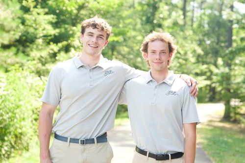Two smiling people in light gray polos standing arm-in-arm outdoors on a sunny green path