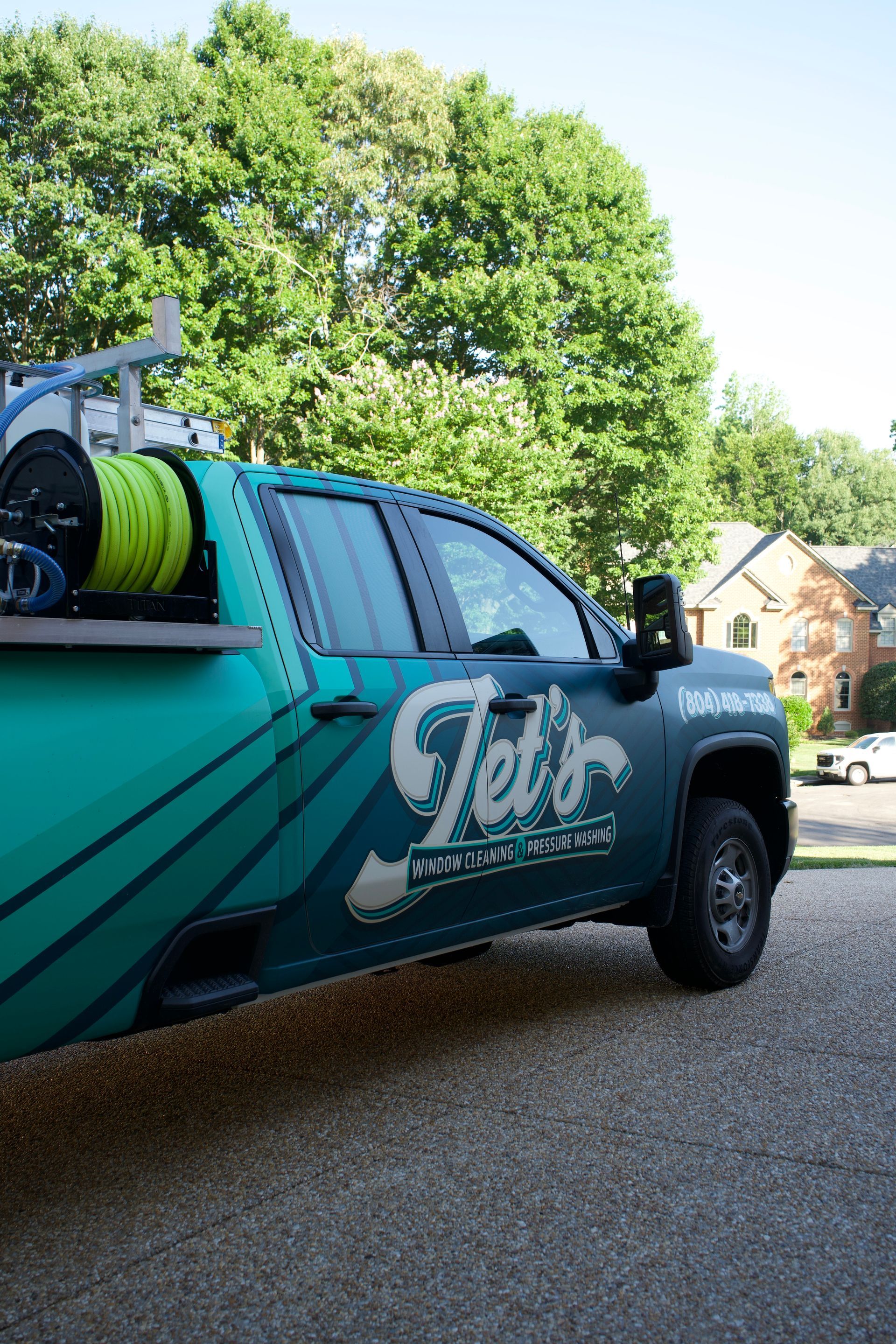 Teal plumbing service truck with hose reels parked in a driveway, trees and a house in the background