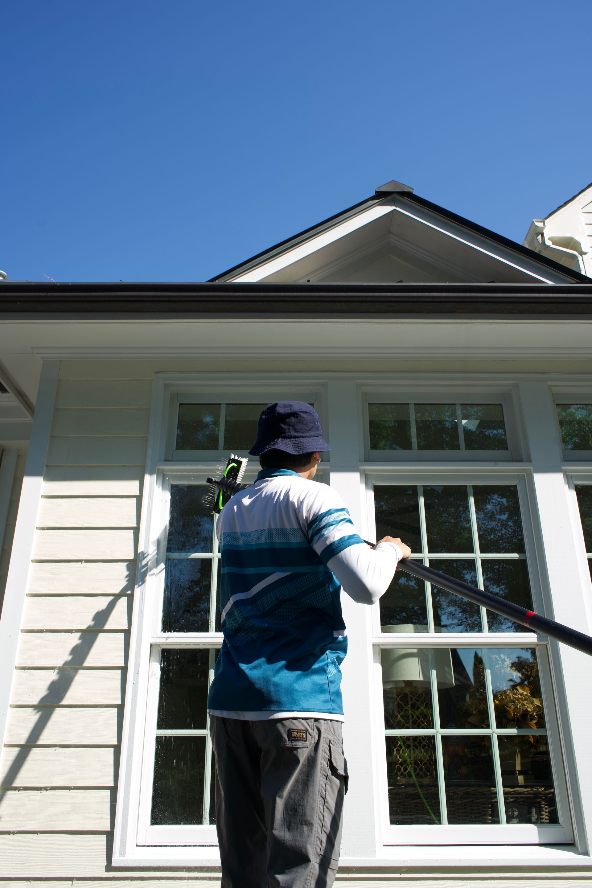 Person painting the exterior window trim of a white house with a roller under a blue sky