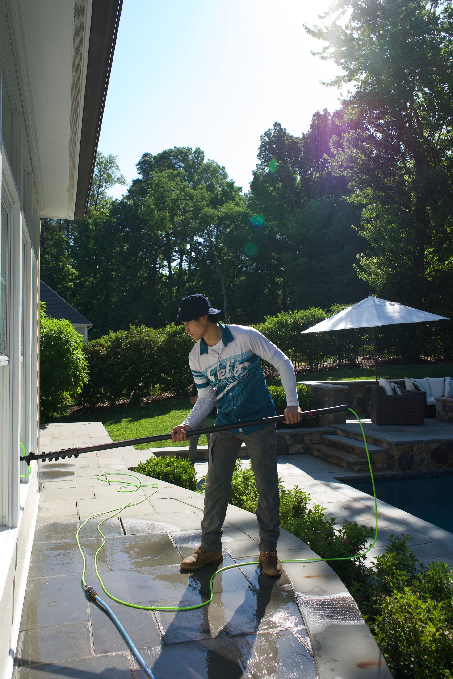 Person washing a patio with a hose beside a house, in bright sunlight.