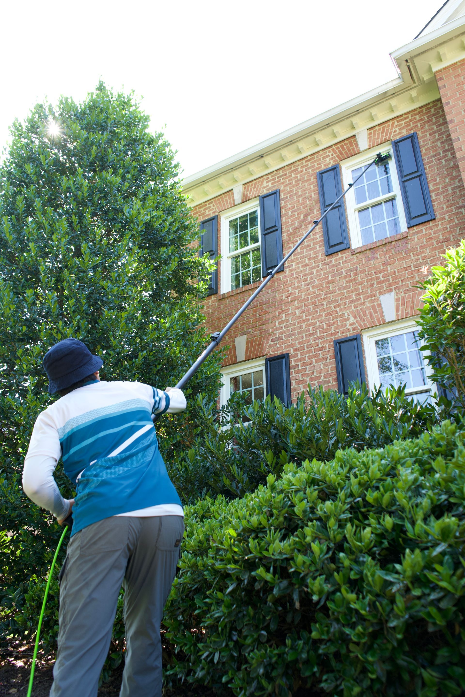 Person spraying a brick house with a long-handled sprayer near bushes