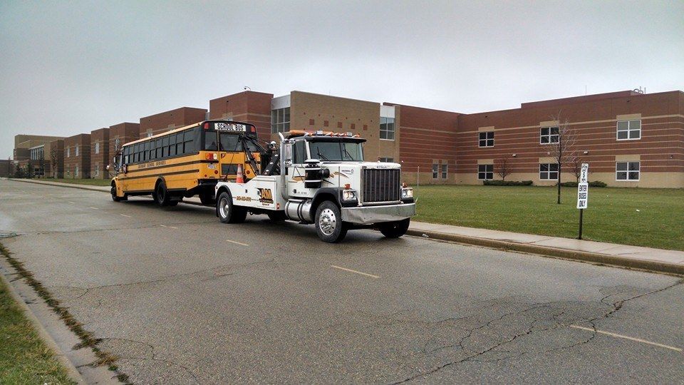 Tow truck towing a yellow school bus in front of a brick building on a cloudy day.