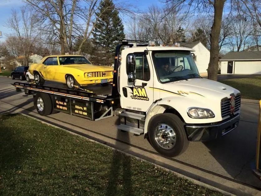 Yellow classic car on a flatbed tow truck. White truck, grassy area, sunny day.
