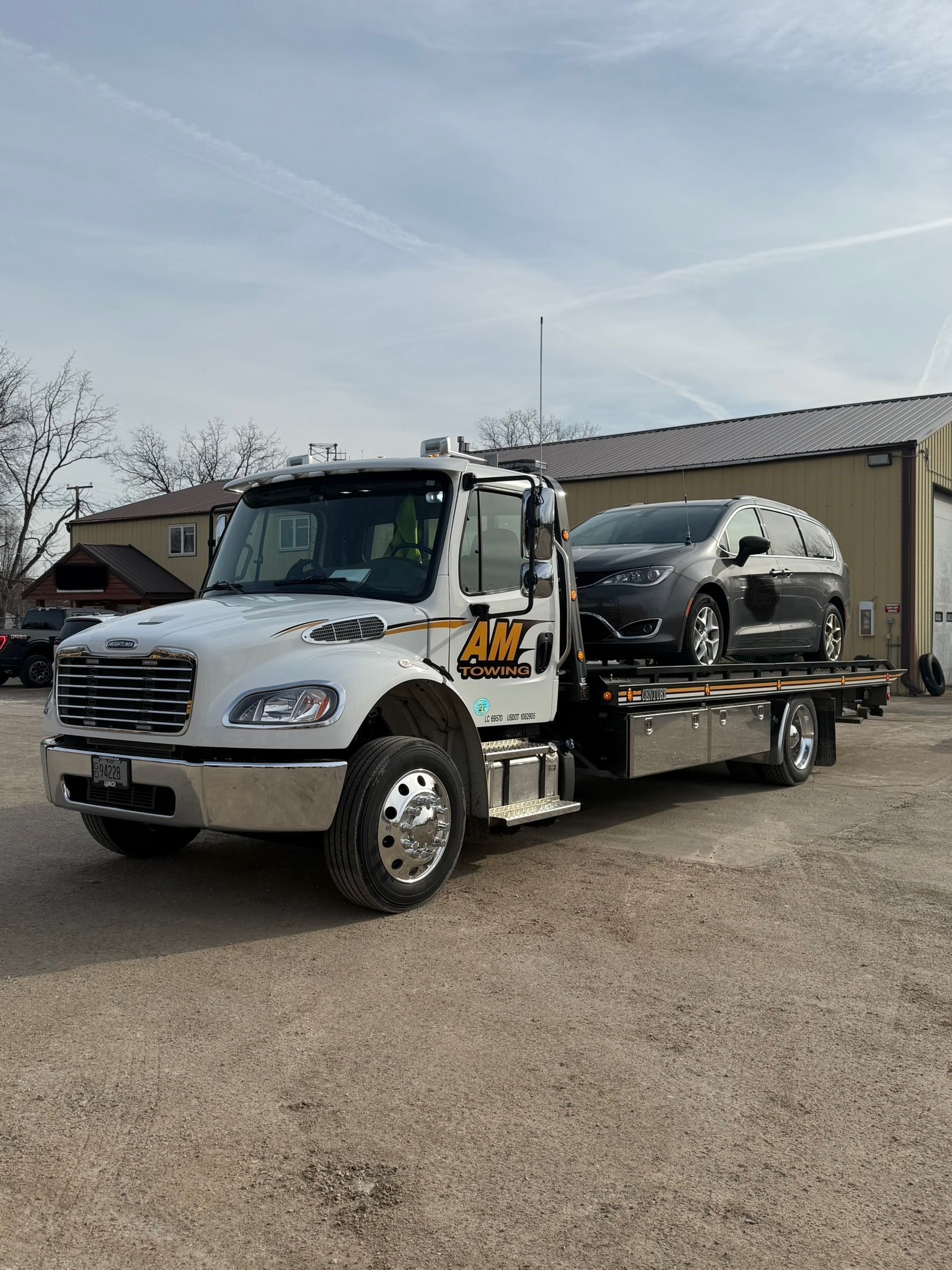 White tow truck with car on its bed, parked on gravel in front of a building on a partly cloudy day.