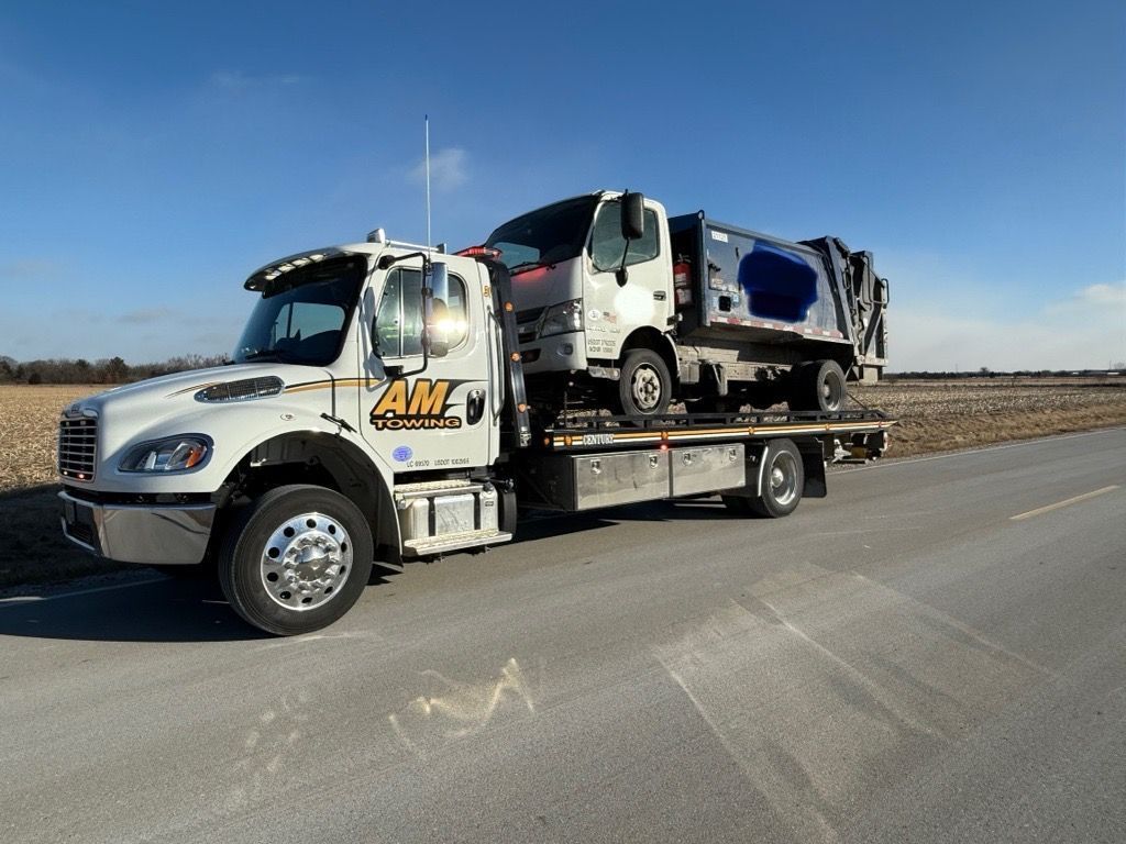 Tow truck with a garbage truck loaded on its flatbed, parked on a road. Bright, sunny day.