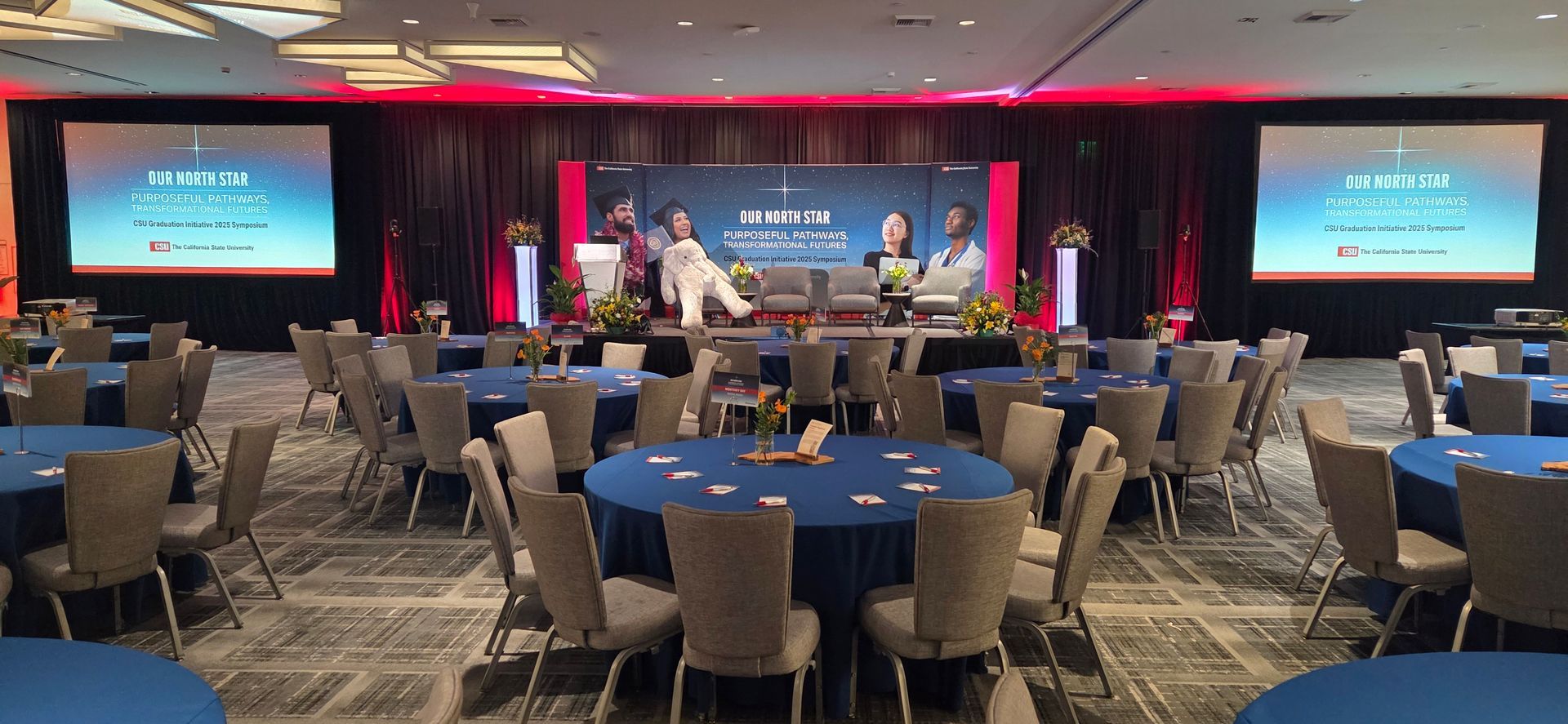 Conference room with blue tables and chairs, panel on stage with presentation screens.