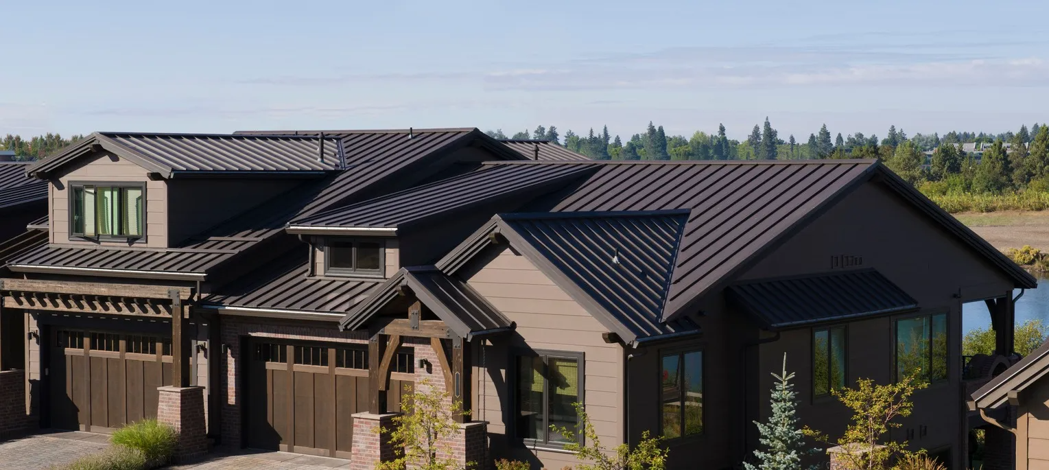 Brown house with a dark metal roof, surrounded by trees, under a blue sky.
